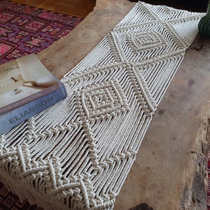 May include: A white macrame table runner with a geometric diamond pattern. The runner is made of natural cotton cord and has a fringe on the ends. The runner is laying on a wooden table with a book on top.