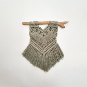 Peut inclure: Une suspension murale en macramé en forme de cœur avec des franges, réalisée avec du fil de couleur vert clair et une branche de bois naturel.