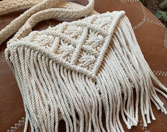 May include: A white crocheted bag with a triangular design and long fringe. The bag has a long strap and is made of thick rope.