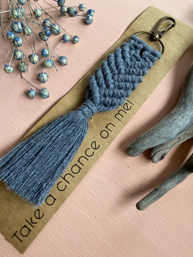 May include: Grey macrame keychain with a tassel and a silver key ring. The keychain has the text "Take a chance on me!" written on it.