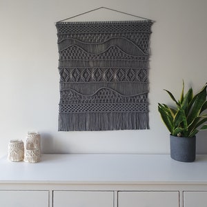 May include: Gray macrame wall hanging with a geometric pattern. The hanging has a fringe at the bottom and is hung on a white wall above a white dresser with three drawers.