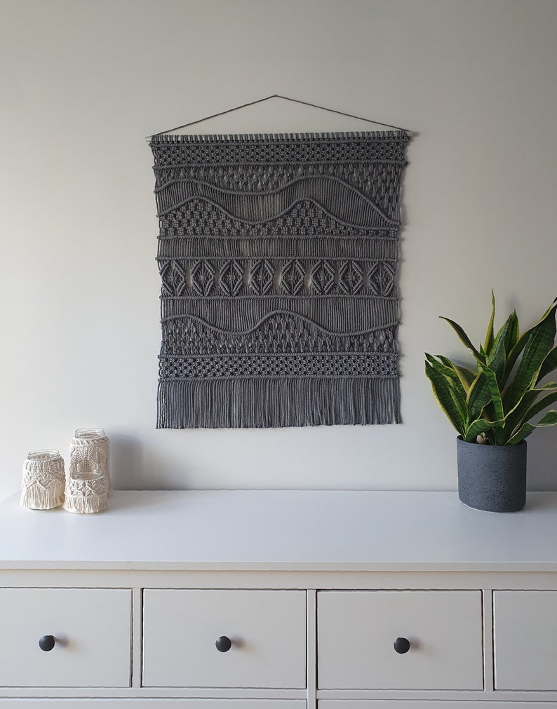 May include: Gray macrame wall hanging with a geometric pattern. The hanging has a fringe at the bottom and is hung on a white wall above a white dresser with three drawers.