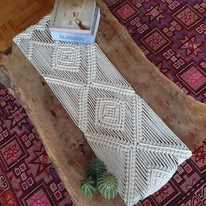 May include: A white macrame table runner with a diamond pattern on a wooden surface. The runner is made of thick cotton cord and has a fringed edge. The runner is on top of a colorful rug.
