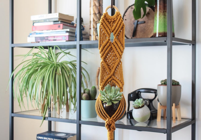 K&ouml;nnte beinhalten: Ein senfgelber Makramee-Pflanzenh&auml;nger mit einem Holzring oben. Eine kleine Sukkulente befindet sich in einem schwarzen Topf im H&auml;nger.