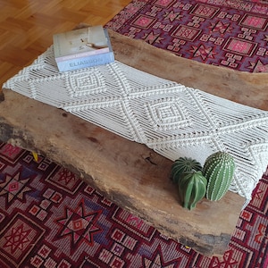 May include: A white macrame table runner with a diamond pattern sits on a wooden table with a natural finish. The table is on top of a red and purple patterned rug. There are three small green succulent plants on the table.
