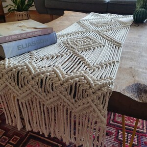 May include: A white crocheted table runner with a diamond pattern and fringe. The runner is on a wooden table with books on top.