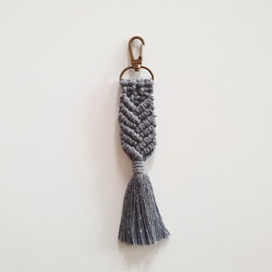 May include: A grey macrame keychain with a tassel. The keychain has a metal ring and clasp.