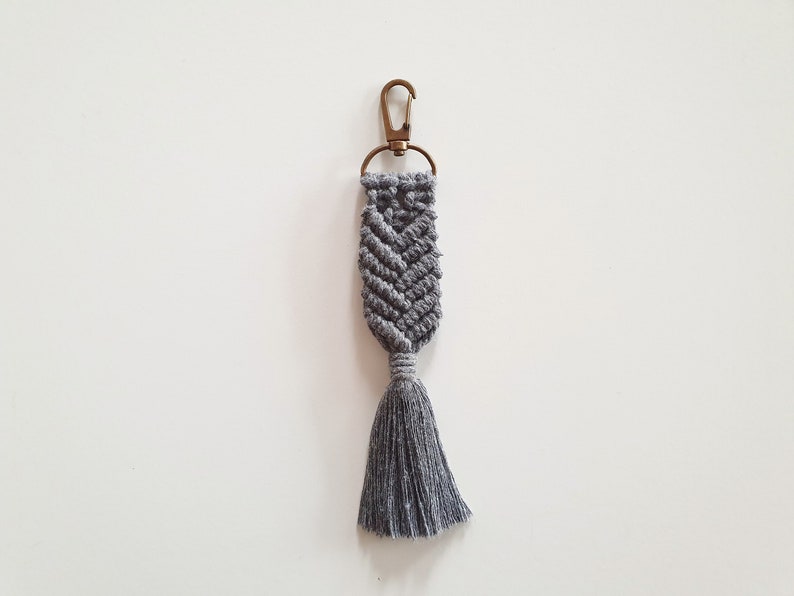 May include: A grey macrame keychain with a tassel. The keychain has a metal ring and clasp.