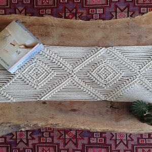 May include: A white macrame wall hanging with a geometric diamond pattern. The macrame is hanging on a wooden surface with a book titled "ELLIASSEN INTERIOR ARCHITECTURE" on top of it.