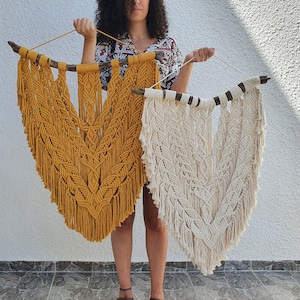 以下が含まれることがあります： 2つのマクラメの壁掛け。1つはマスタードイエロー、もう1つはオフホワイトです。どちらも天然の木の枝から吊り下げられています。マクラメは、複雑な結び目とフリンジを備えた中央のシェブロンデザインが特徴です。