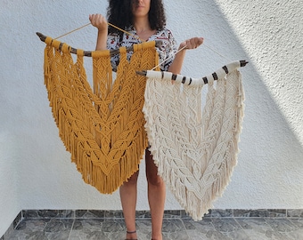 May include: Two macrame wall hangings. One is mustard yellow, the other is off-white. Both are suspended from a natural wooden branch. The macrame features a central chevron design with intricate knotting and fringe.