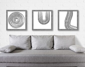 May include: Three framed black and white abstract line art prints with a gray frame. The prints are hung on a white brick wall above a gray couch.