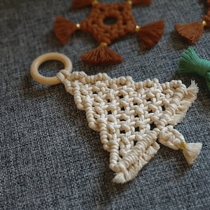May include: A white macrame Christmas tree ornament with a wooden ring at the top. The ornament is hanging on a grey textured surface.