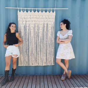 May include: Two people stand in front of a macrame wall hanging. The wall hanging is made of natural fibre rope and has a geometric pattern. The person on the left is wearing a black tank top and denim shorts. The person on the right is wearing a white floral dress.