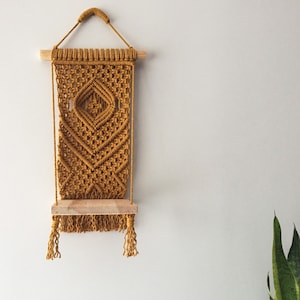 May include: A mustard yellow macrame wall hanging with a wooden shelf. The macrame has a diamond pattern and fringe.