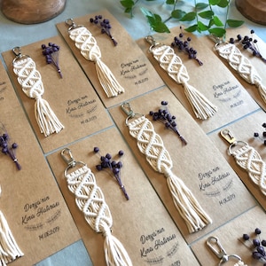 May include: A collection of handmade macrame keychains with a tassel on a brown card with the text "Derya'nin Kina Hatırası 14.06.2019" printed on it. The keychains are decorated with small sprigs of dried purple flowers.