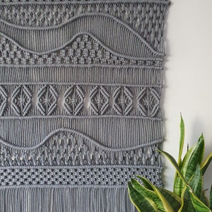 May include: Grey macrame wall hanging with a geometric pattern and fringe. The hanging is made with thick cotton cord and has a woven design.