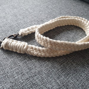 May include: A white cotton camera strap with a brass clasp. The strap is braided and has a simple design.
