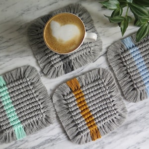 May include: Four gray crocheted coasters with fringe edges. Each coaster has a different colored stripe in the center: green, blue, yellow, and gray. One coaster has a cup of coffee on top.