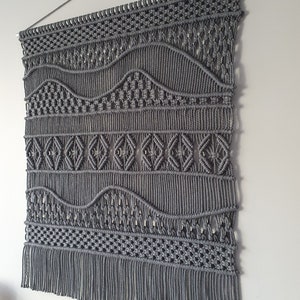 May include: Grey macrame wall hanging with a geometric pattern and fringe. The hanging is made with thick cotton cord and has a woven design.