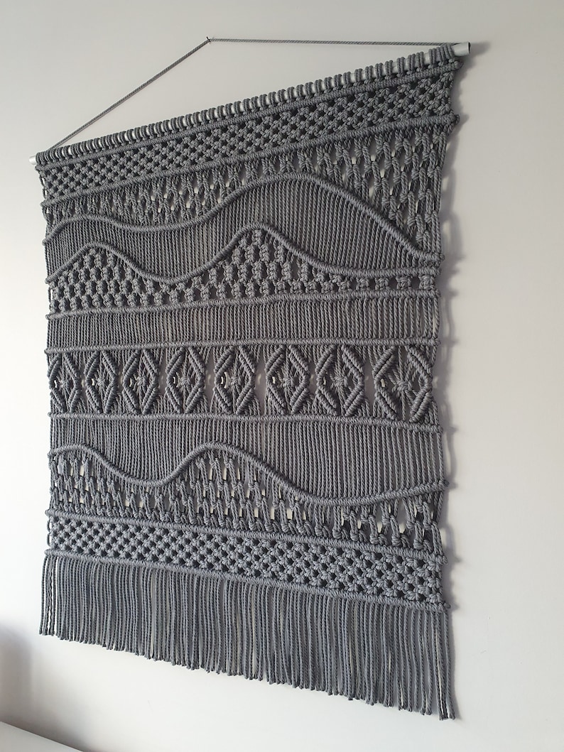 May include: Gray macrame wall hanging with a geometric pattern and fringe. The hanging is made with thick cotton cord and has a woven design.