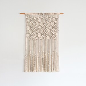 Puede incluir: Un tapiz de macramé blanco con un patrón geométrico y flecos. El tapiz está sujeto a una varilla de madera.