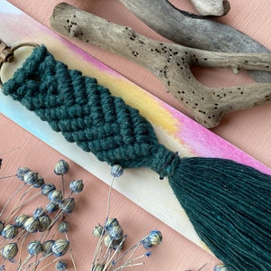 May include: A dark green macrame keychain with a tassel. The keychain is attached to a gold key ring. The keychain is displayed on a wooden surface with a light pink background.