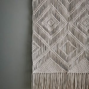 May include: A beige macrame wall hanging with a geometric pattern and fringe.