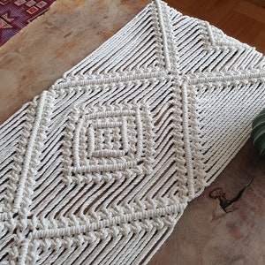 May include: A white macrame table runner with a diamond pattern. The runner is made of thick cotton cord and has a natural, rustic look.