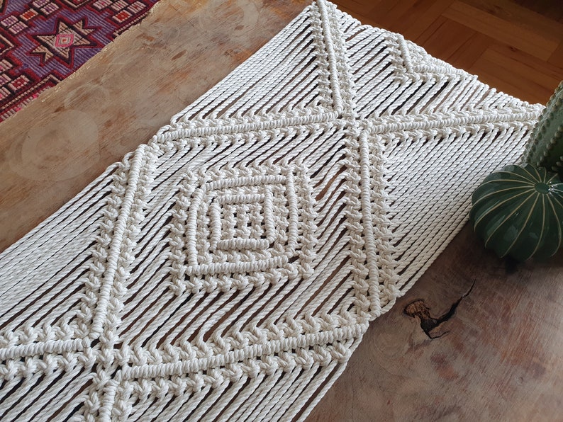May include: A white macrame table runner with a diamond pattern. The runner is made of thick cotton cord and has a natural, rustic look.