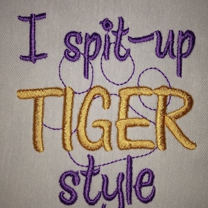 May include: Embroidered design on a white fabric background. The design reads "I spit up tiger style" in purple and gold thread.