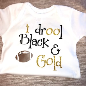 May include: White baby onesie with black text that reads "I drool Black & Gold" with a brown football graphic.