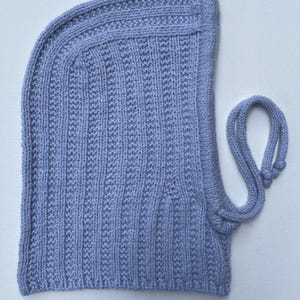 May include: A light periwinkle-colored knitted balaclava with a ribbed texture. The hood features a vertical ribbed pattern and a drawstring closure. The knitted hat is made of soft yarn.