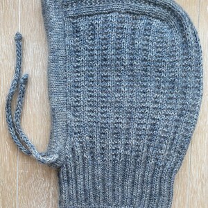 May include: A gray knitted balaclava with a textured, ribbed pattern. The hood features a face opening and attached ties. The balaclava is made of a soft, warm material, perfect for cold weather.