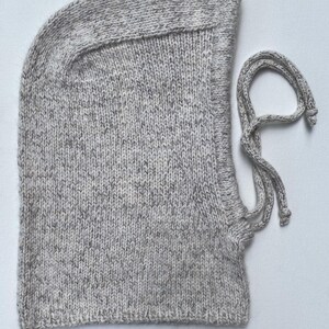May include: A light gray knitted balaclava with a drawstring closure. The hood is made of a speckled yarn, and the drawstring is a matching color. The balaclava is designed to cover the head and neck.