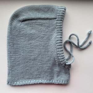 May include: A light blue knitted balaclava with a drawstring closure. The balaclava is made of a soft, textured yarn and has a ribbed edge around the face opening. The drawstring is knotted at the ends.