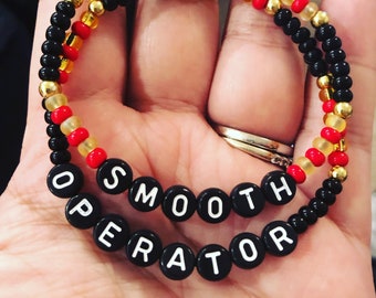 Puede incluir: Un brazalete de cuentas negras con cuentas rojas y doradas. El brazalete tiene cuentas de letras negras que deletrean "SMOOTH OPERATOR".
