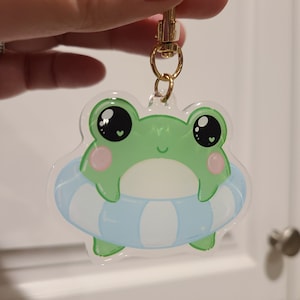 May include: A green frog with big eyes and a pink blush is sitting in a blue and white striped inflatable ring. The frog is attached to a gold keychain.