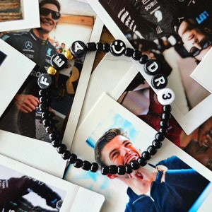 Puede incluir: Un brazalete de cuentas negras con cuentas de letras doradas que deletrean "FEAR DR3" y una cuenta sonriente. El brazalete está sobre una pila de fotos Polaroid.