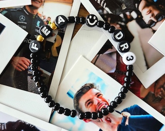 May include: A black beaded bracelet with gold letter beads spelling "FEAR DR3" and a smiley face bead. The bracelet is lying on top of a stack of Polaroid photos.