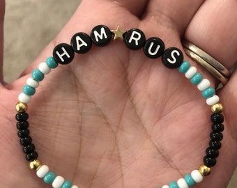 May include: A black and turquoise beaded bracelet with gold beads and black letter beads spelling out the name "HAMRUS" with a gold star bead between the "M" and "R".