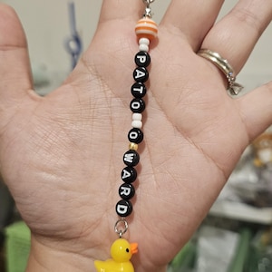 Puede incluir: Un dije de cuentas con la palabra "PATO OWARD" escrita con cuentas de letras negras. El dije presenta un pequeño pato de goma amarillo y una cuenta a rayas naranja y blanca. El dije está unido a un cordón negro.
