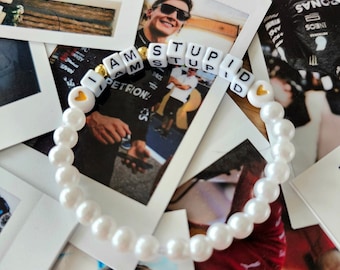 May include: A white pearl bracelet with gold heart charms and white letter beads spelling "I AM STUPID". The bracelet is photographed on top of a stack of Polaroid photos.