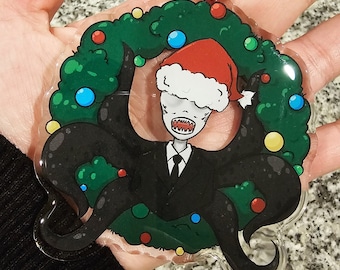 May include: A clear plastic Christmas ornament featuring a cartoon Slender Man wearing a Santa hat and a black suit. He is surrounded by a green wreath with red, yellow, blue, and green ornaments.