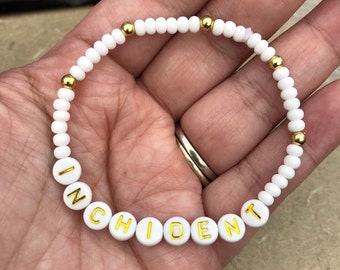 May include: White beaded bracelet with gold beads and gold lettering spelling out the word "INCIDENT".