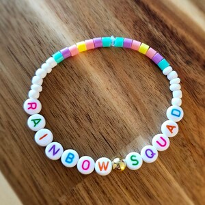 May include: A colourful beaded bracelet spelling "RAINBOW SQUAD". The bracelet features white, pastel, and gold beads. The letter beads are in various colours. The bracelet is displayed on a wooden surface. A fun and vibrant accessory.
