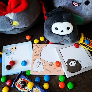 May include: A set of three blank notepads featuring different Halloween-themed illustrations. The illustrations include a ghost, a skeleton, and a cat. The notepads are surrounded by colourful sweets and plush toys.