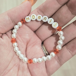 Peut inclure: Bracelet en perles blanches avec le nom "NORRIS" écrit en lettres dorées sur des perles blanches. Le bracelet comporte également de petites perles orange.