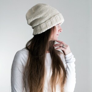May include: A woman with long brown hair wearing a white knitted beanie hat and a white long-sleeved shirt.