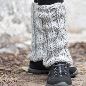 May include: Gray knitted leg warmers with a cable knit pattern. The leg warmers are worn over black sneakers.